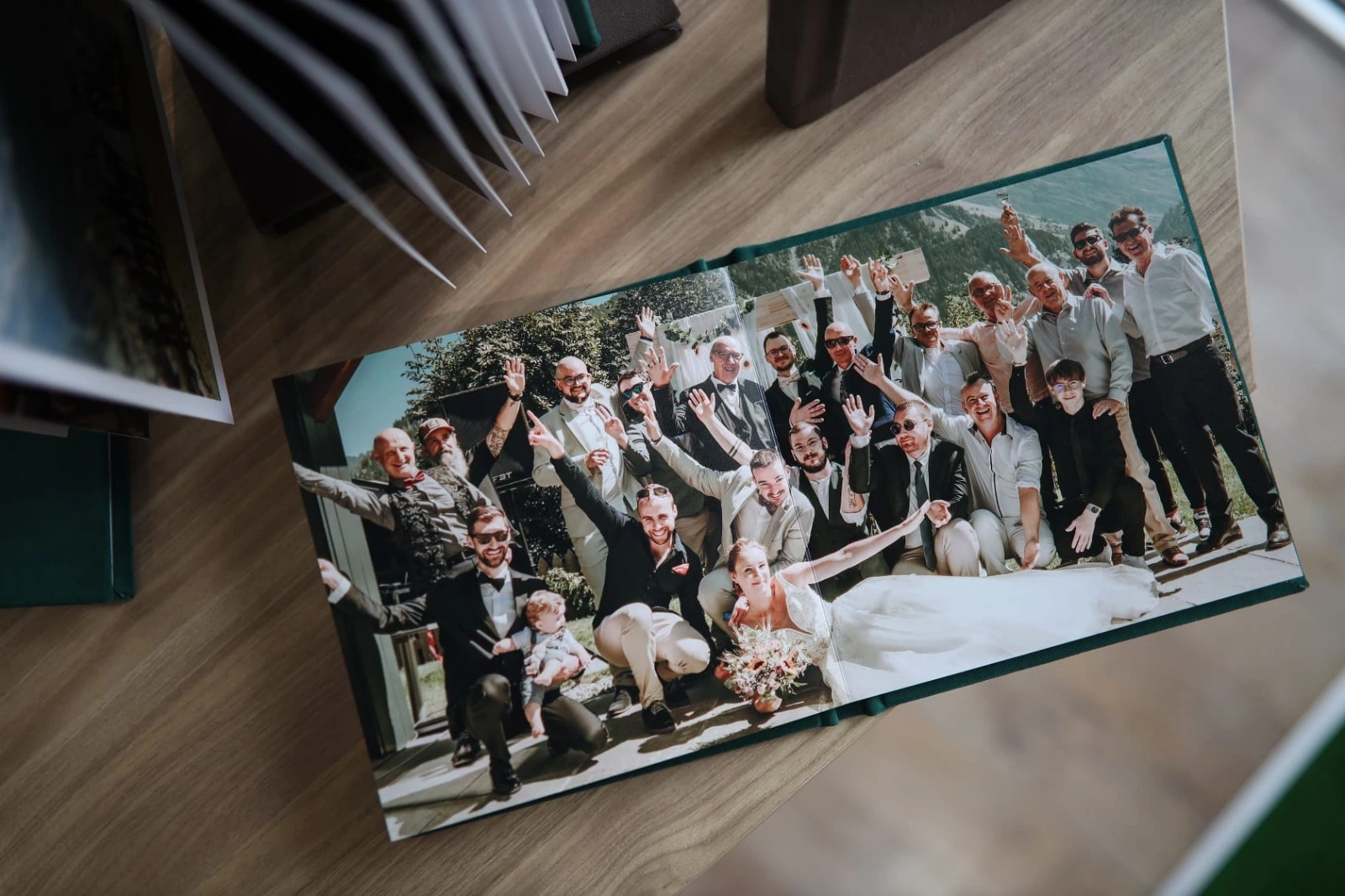 A wedding photo album opened to a page displaying a joyful group wedding photo. The bride, groom, and groomsmen are posing together with smiles and raised hands in an outdoor setting with greenery and mountains in the background. The album is placed on a wooden table, surrounded by other albums.