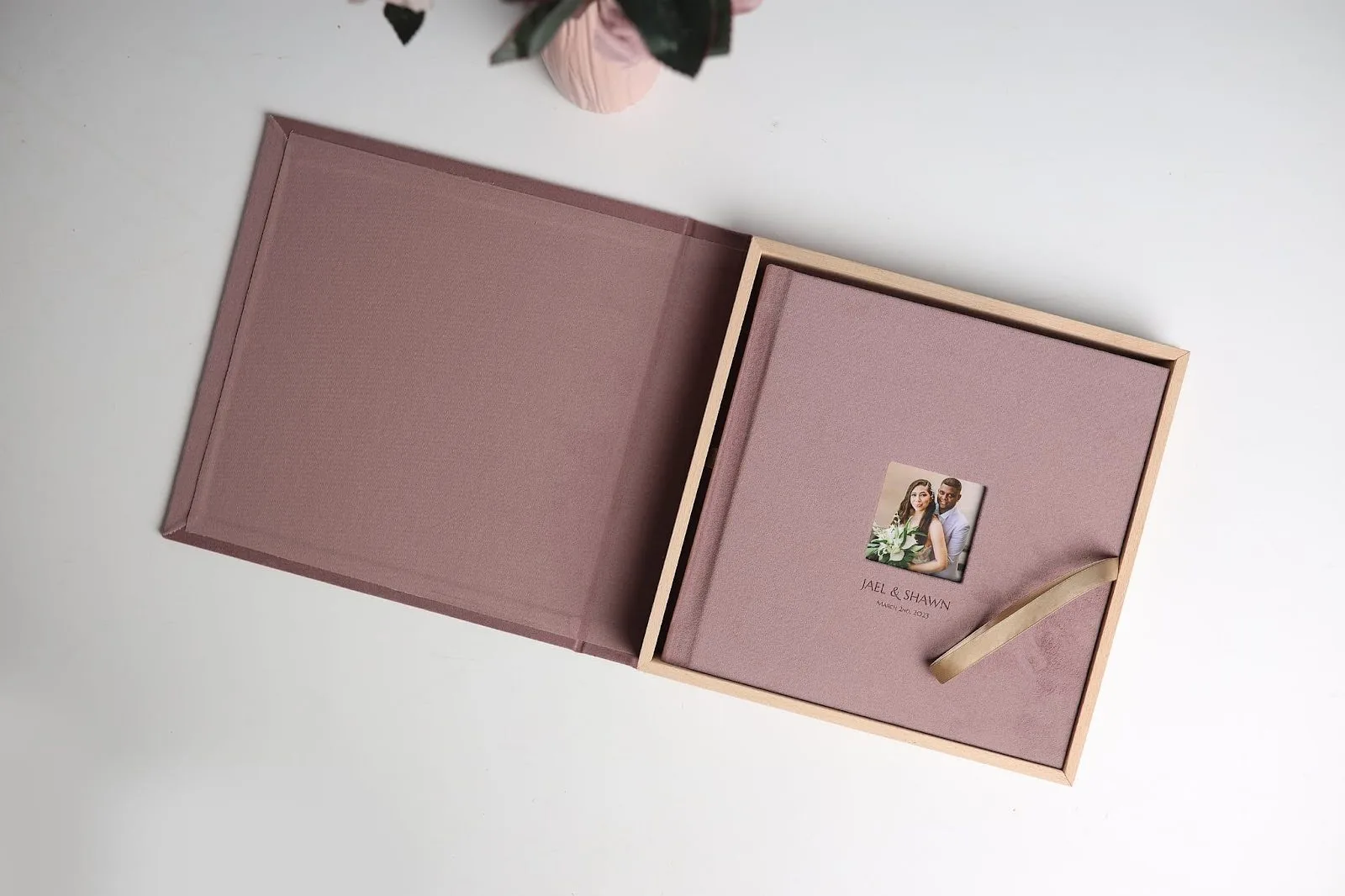 A wedding photo album in a mauve fabric-covered box. The cover has a small picture of a couple with the text "JAEL & SHAWN, October 20, 2023." A ribbon secures the album inside the wooden-framed box, placed on a white surface with a plant in the background.