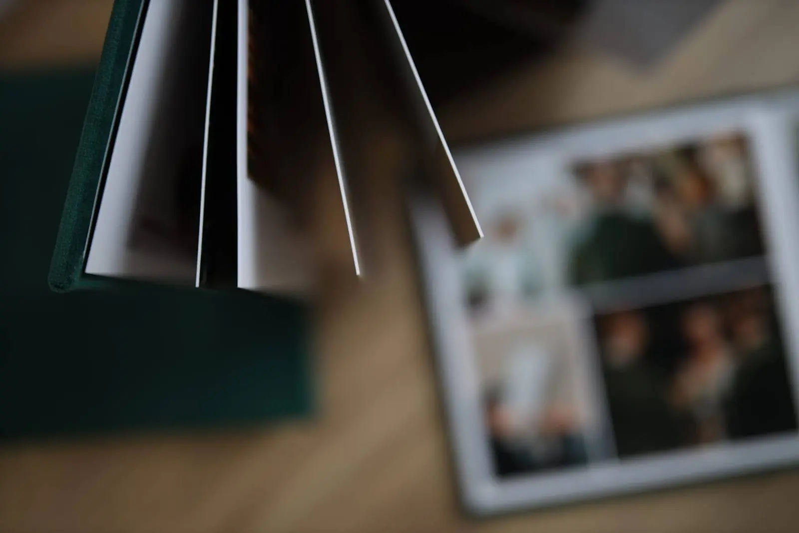 Close-up of an open photo album with thick, high-quality pages, placed on a table. A second blurred photo album with wedding or event images is visible in the background.
