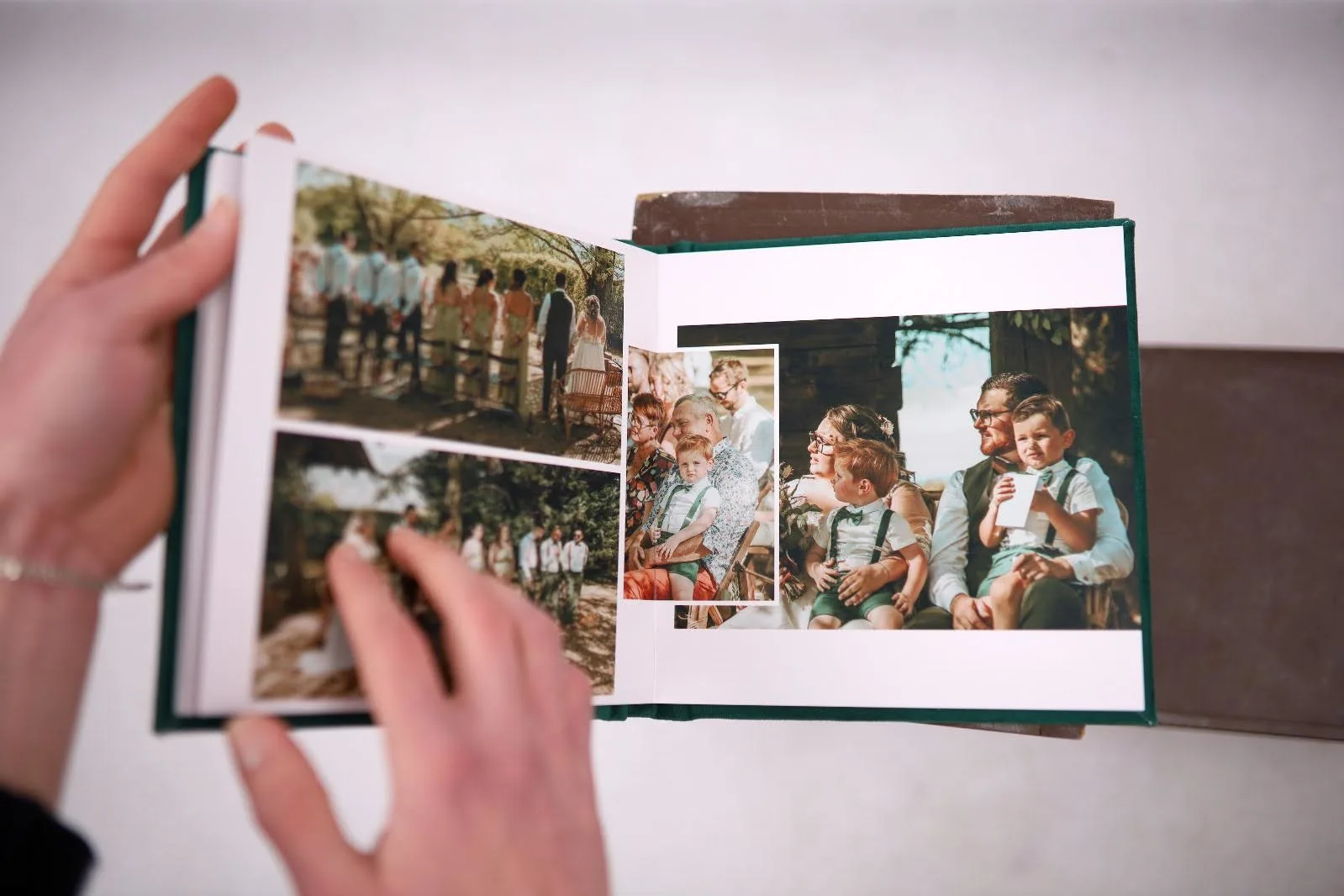 A person is holding an open wedding photo album, displaying pictures of an outdoor ceremony and guests. The pages feature images of a wedding party, seated attendees, and children dressed in formal outfits. The album has a green cover and rests on a brown surface in the background.