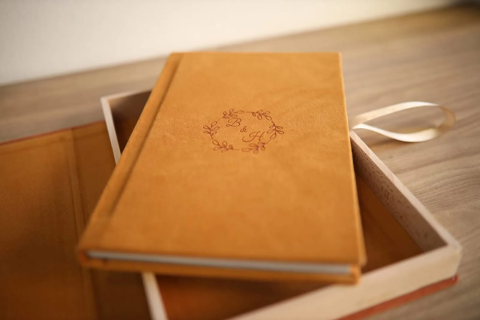 A yellow photo album with 'B & H' on the cover, placed in an open wooden box with a ribbon, sitting on a wooden table.