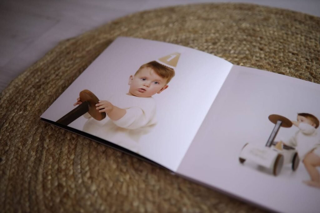 A close-up of a photo book showing a child wearing a birthday hat, holding a toy. The child is on a woven rug, and the photo shows them in a playful, innocent pose.