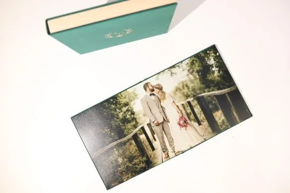 A wedding photo album with a green cover, featuring a photo of the bride and groom standing on a bridge, holding hands and kissing. The album is placed on a white surface.