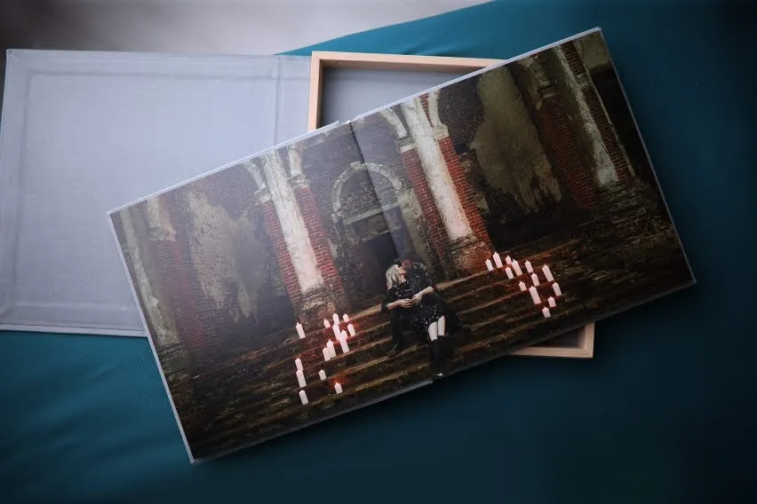 A wedding photo album open to a photo of a couple sitting on steps in a grand, old building surrounded by candles. The album is placed on a teal surface with a beige box beside it.
