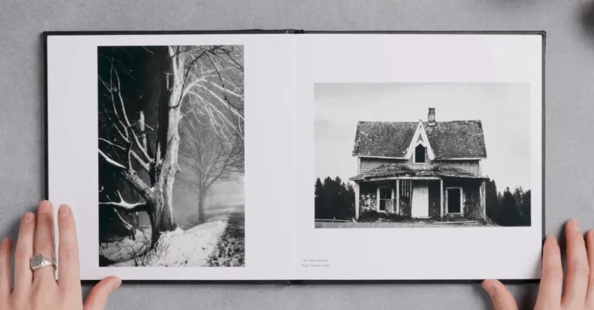 A photo album open to two black-and-white images: one of a tree-lined path in the snow, and the other of an old, abandoned house. A hand rests on the album's edge.