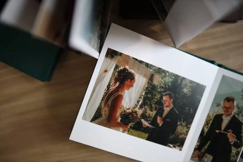 A close-up of a wedding photo album showing a bride and groom during their ceremony, with the bride holding flowers and the groom reading his vows. The photo is taken in a sunny outdoor setting.