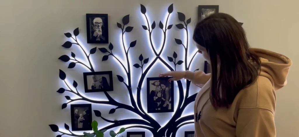 A woman pointing at a framed photo on a decorative family tree wall display with glowing backlight.