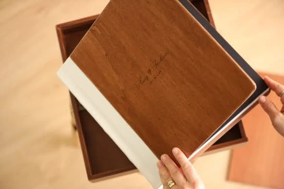 A person holding a wooden wedding photo album with engraved names and dates on the cover.