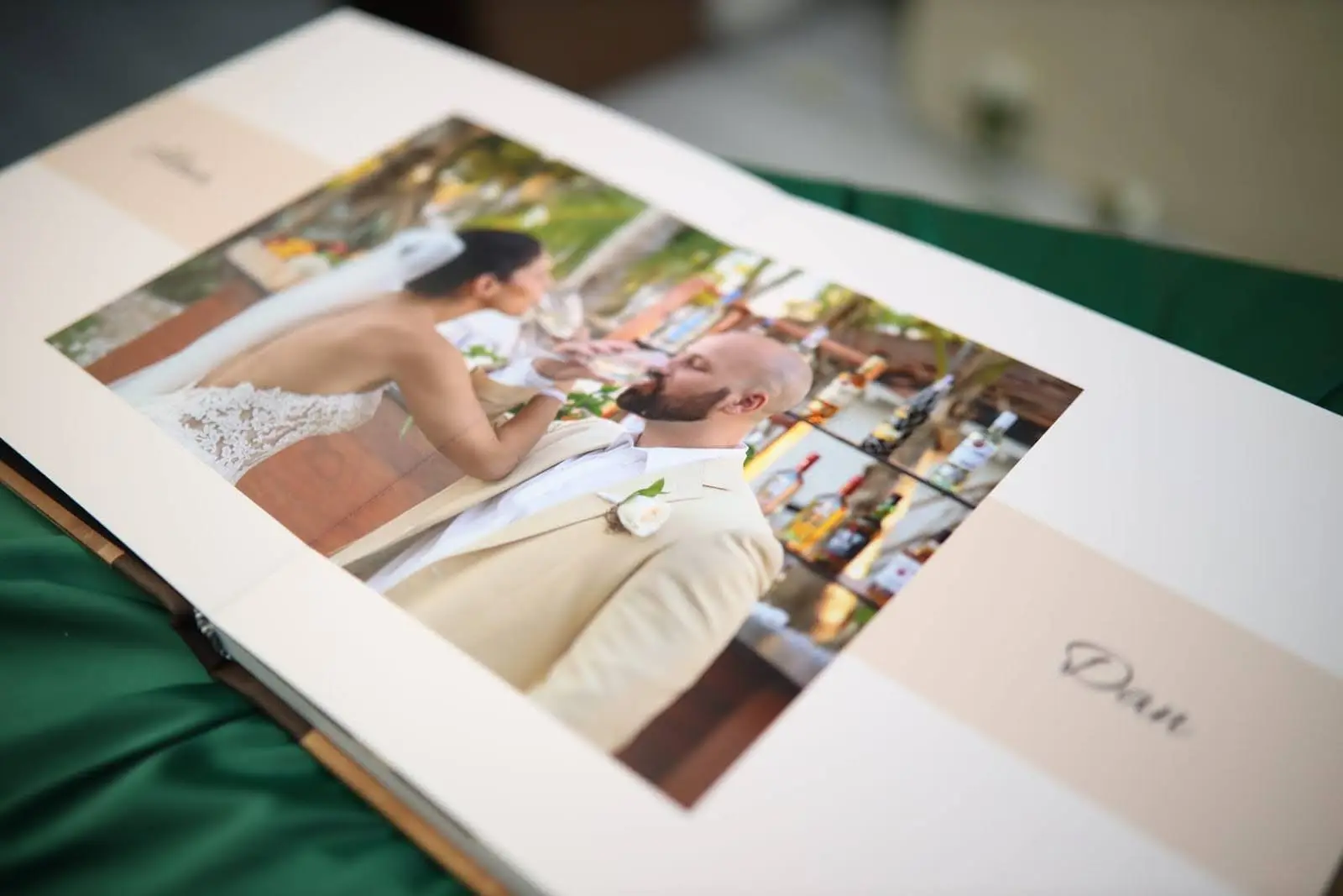 Open wedding photo album showing a bride and groom sharing a toast at an outdoor bar. The page is elegantly designed with soft beige accents and the groom’s name, “Dan,” printed on the right.
